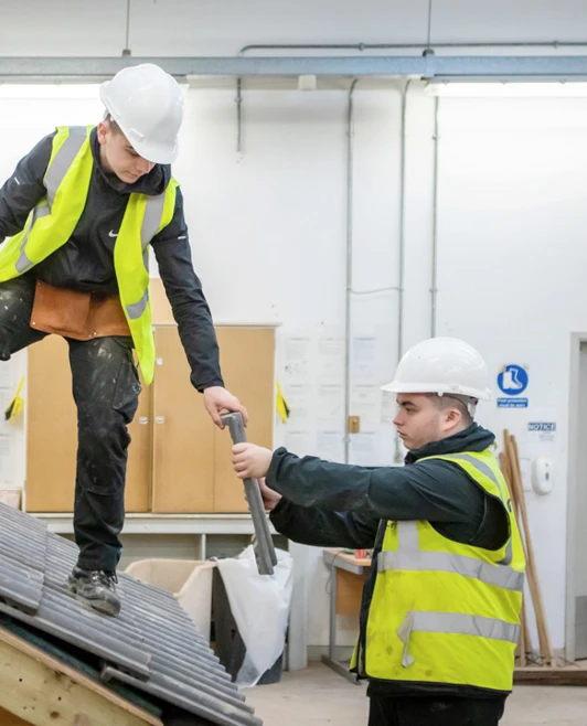A roofing student climbing a wooden training structure while receiving a roof tile from a fellow student during practical training. A roofing student climbing a wooden training structure while receiving a roof tile from a fellow student during practical training.